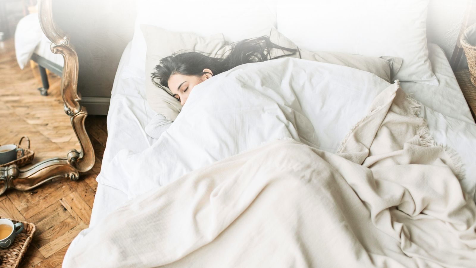 Woman with long brown hair snuggled into cream linen bedding on a wooden floor, creating a peaceful evening atmosphere