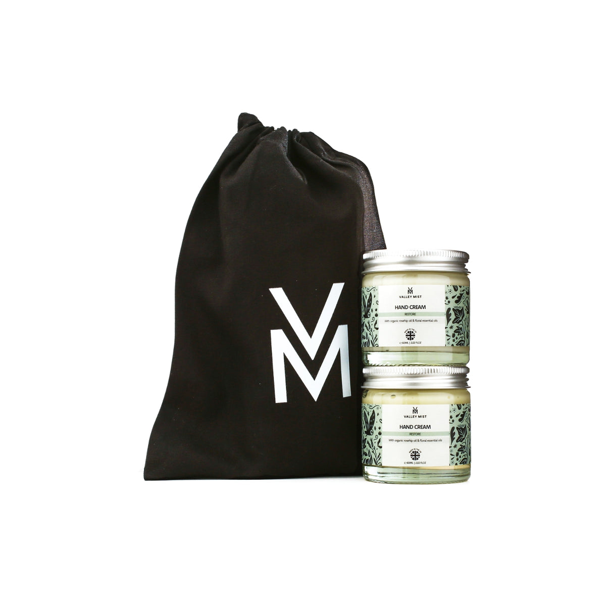 Two small glass jar candles with silver lids and white labels are displayed next to a black drawstring bag with a large white 'VM' logo, on a plain white background.
