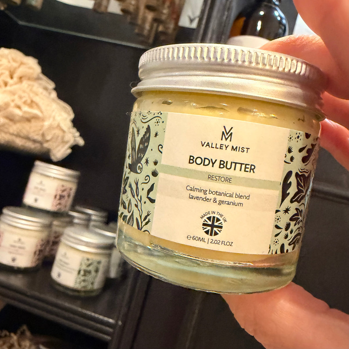 A hand holding a glass jar of Valley Mist Restore Body Butter, showing the cream-colored butter and the label, with other jars blurred in the background on a shelf.
