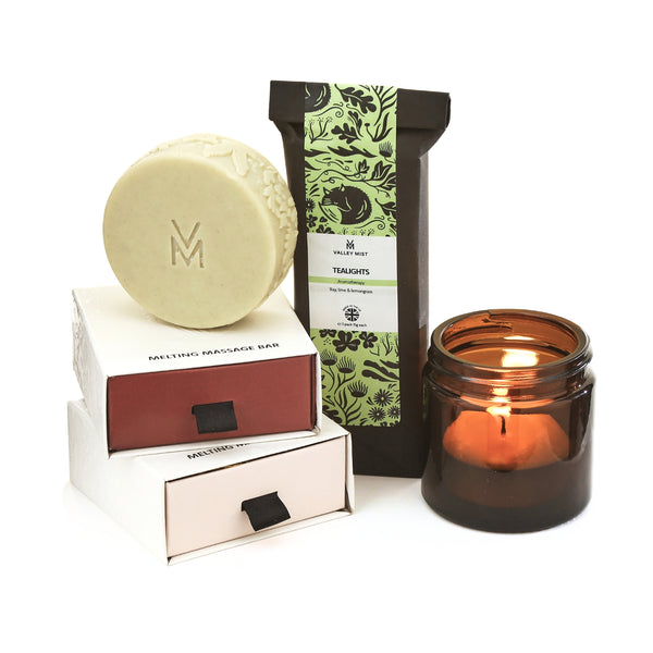 Massage  bars and a tea-light scented gift set from Valley Mist, featuring a round beige massage bar, a black pouch of tea-light scented candles with green floral patterns, and an amber glass jar candle with a lit flame, arranged on a white background.