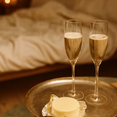 Organic cocoa butter massage bar on a tray with two glasses of champagne in a softly lit romantic bedroom setting.